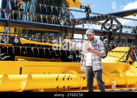 Un agricoltore maschio si trova accanto a una nuova mietitrebbia presso un concessionario di attrezzature agricole, leggendo un contratto di leasing su un tablet digitale. Foto Stock