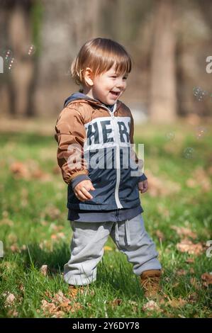 In un parco vivace pieno di luce soffusa, un bambino allegro ride e gioca con gioia, circondato da scintillanti bolle e colorate foglie autunnali Foto Stock