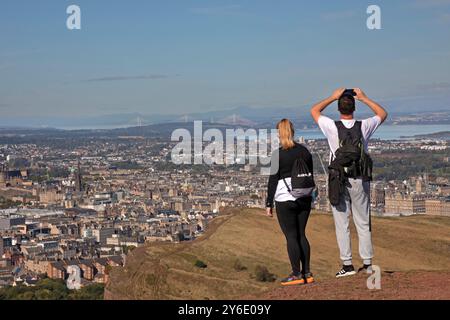 Arthur's Seat summit, Edimburgo, Scozia, Regno Unito. 25 settembre 2024. Affollato sulla cima dell'estinto vulcano nel Holy Rood Park. Persone di tutte le età hanno fatto il viaggio lungo i vari sentieri per raggiungere il punto di vista della capitale scozzese. Temperatura 8 gradi a metà mattina, fino a 11 gradi a metà pomeriggio. Nella foto: Una coppia ammira la vista sul centro della città verso i Forth Bridges e la costa di Fife sullo sfondo. Crediti: Archwhite/alamy notizie in diretta. Foto Stock