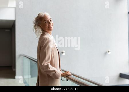 Donna sorridente in abiti da lavoro in piedi accanto a una ringhiera in un moderno edificio per uffici, spazio per copie Foto Stock