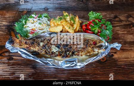 Vista dall'alto ravvicinata della trota appena pescata con salsa, patatine fritte e insalate fresche e succose su un tavolo rustico in legno Foto Stock