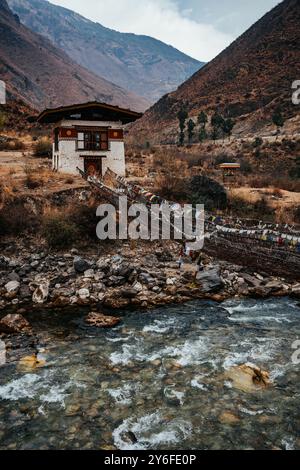 Una tradizionale casa bhutanese con un ponte sospeso adornato con bandiere di preghiera sopra un fiume in un paesaggio montuoso. Foto Stock