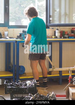 Il meccanico che indossa una maglietta verde sta serrando una parte della bicicletta in una morsa da banco nella sua officina, utilizzando una chiave e indossando guanti Foto Stock