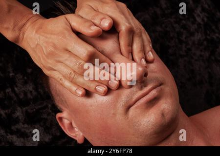 Primo piano dell'uomo che riceve un massaggio facciale, le mani del massaggiatore massaggiano la pelle del viso su sfondo scuro Foto Stock