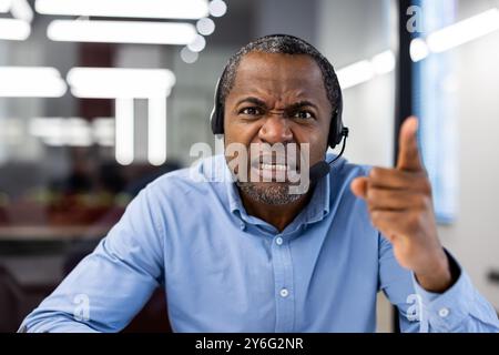 Uomo frustrato e arrabbiato con la camicia blu e la cuffia che punta il dito mentre parla in ufficio. Le emozioni espressive durante la conversazione con le videochiamate creano tensione e stress sul posto di lavoro. Foto Stock