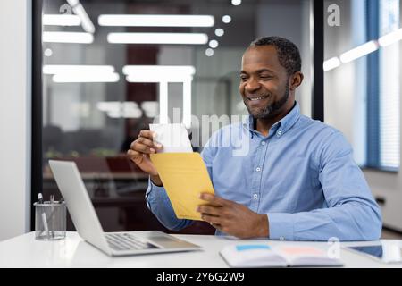 Un lavoratore d'ufficio apre la busta della posta alla scrivania, sorridendo alle buone notizie. Ambiente di lavoro con notebook, che mostra produttività, successo e soddisfazione in ufficio. Rappresenta un risultato professionale. Foto Stock