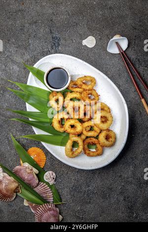 Cibo, anelli di calamaro fritti su piatto bianco decorato con foglie tropicali, sfondo grigio in cemento, vista dall'alto Foto Stock