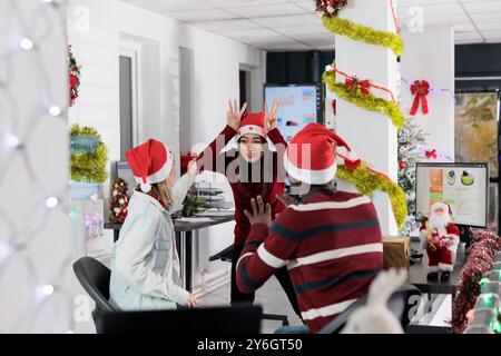 Dipendente asiatico che gioca a sciagole con il personale dell'azienda durante i turni di pausa, imitando le renne durante le festività natalizie. Colleghi che si divertono insieme, si legano in un ufficio decorato con festività Foto Stock