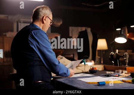 Un'anziana couturier che guarda i disegni di schizzo, si prepara a creare capi sartoriali personalizzati per il cliente. Suitmaker che concettualizza gli abiti seguendo bozze disegnate a mano Foto Stock