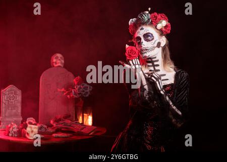 Donna che tiene una rosa rossa, che fa riti indigeni di onorare i parenti morti durante dia de los Muerte. Lady che indossa il trucco del cranio partecipa all'evento Day of the Dead, Messico, in onore del defunto Foto Stock