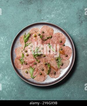 Cibo, carpaccio di manzo marmorizzato con rucola e capperi su piatto grigio, blu legno, vista dall'alto Foto Stock