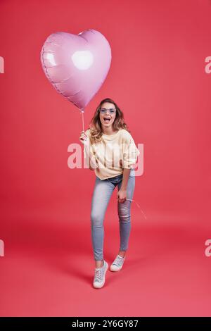 Sentitevi liberi di fare qualsiasi cosa. L'intera lunghezza di una giovane donna sorridente che tiene in mano il palloncino a forma di cuore e urla mentre sta in piedi contro il dorso rosa Foto Stock
