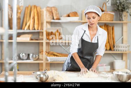 La panettiera femminile si trova al banco di lavoro, impastando e modellando l'impasto per fare pane, croissant e baguette Foto Stock