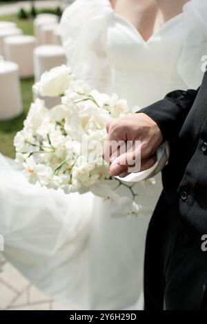 Lo sposo si erge con orgoglio, reggendo un delicato bouquet di fiori bianchi, aggiungendo un tocco di eleganza alla gioiosa cerimonia di nozze all'aperto sotto il bri Foto Stock