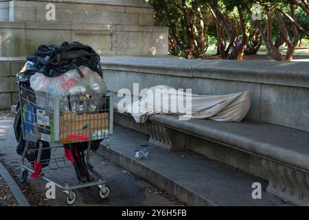 Un senzatetto con il suo carrello pieno di lattine per incassare nelle vicinanze, dorme su una panchina di pietra all'ingresso di Prospect Park durante il pomeriggio a Brooklyn, New York. Foto Stock