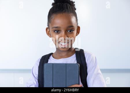 Sorridente ragazza afroamericana che tiene un tablet e indossa uno zaino in classe Foto Stock