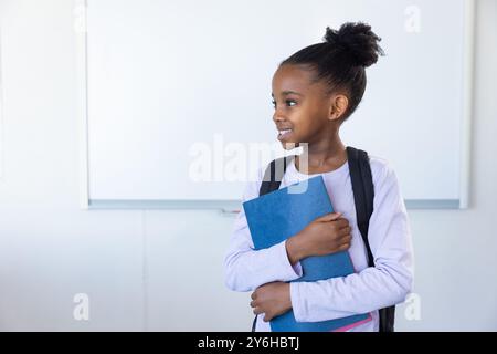 A scuola, una ragazza afroamericana che tiene il quaderno blu e sorride, indossa lo zaino Foto Stock