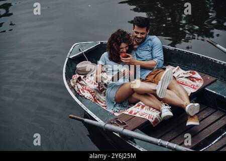 Coppia carina. Bel giovane che dà da mangiare alla sua ragazza con una mela mentre è seduto sulla barca Foto Stock