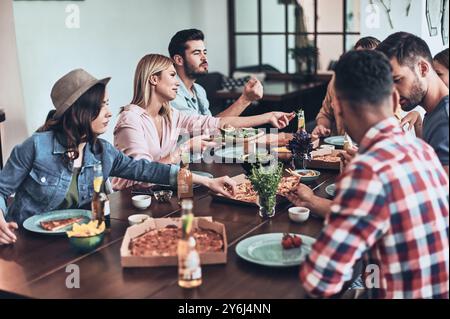 La vita è migliore con gli amici. Gruppo di giovani in abbigliamento casual che mangiano e sorridono mentre fanno una cena in casa Foto Stock