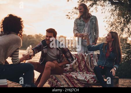 Momento di vera gioia. Gruppo di giovani in abbigliamento casual sorridenti e brindati a vicenda mentre si godono una festa in spiaggia vicino al lago Foto Stock