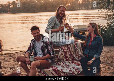 La vita è fantastica! Gruppo di giovani in abbigliamento casual sorridenti e brindati a vicenda mentre si godono una festa in spiaggia vicino al lago Foto Stock