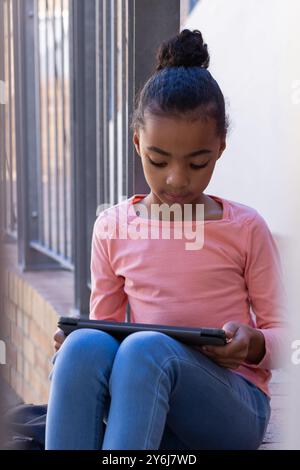 Utilizzando un tablet, una ragazza seduta e che studia nei corridoi della scuola, concentrata sullo schermo Foto Stock