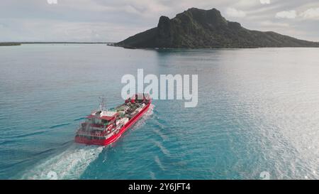 La nave da carico rossa trasporta la portarinfuse in luoghi esotici remoti, Maupiti, Polinesia francese. Grande imbarcazione che naviga sull'isola tropicale d'acqua turchese, giorno di sole. Esportazione e importazione. Vista aerea, volo con droni Foto Stock