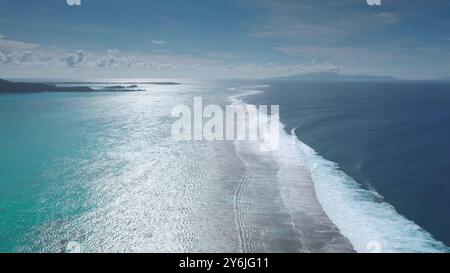 Acqua di mare turchese, cielo soleggiato blu, onde dolci che si infrangono sulla costa tropicale dell'isola, paradiso idilliaco e sereno. Motu della barriera corallina di colore diverso. Paesaggio naturale di bellezza. Colpo statico del drone Foto Stock