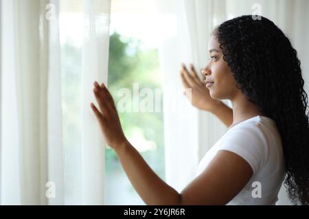 Donna nera seria che apre tende e guarda attraverso una finestra a casa Foto Stock