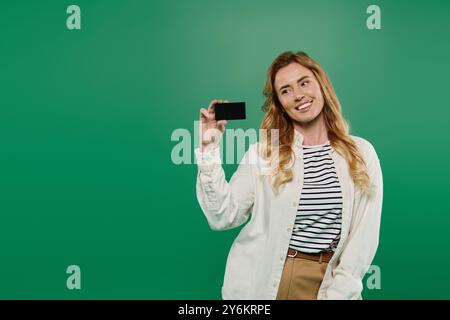 Una donna con i capelli ricci sorride brillantemente mentre tiene in mano una carta, vestita casualmente su uno sfondo verde. Foto Stock