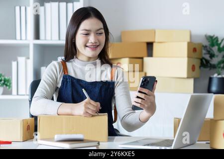 Giovane donna che gestisce il business online dall'ufficio domestico con smartphone e laptop circondato da scatole di spedizione Foto Stock