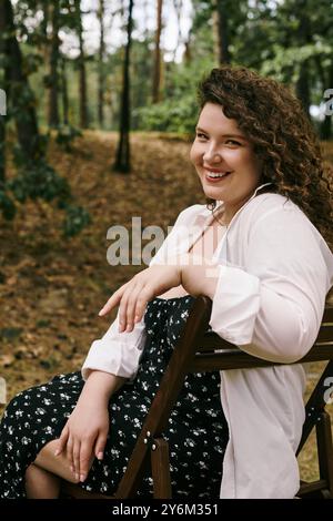 Una donna vivace e di grandi dimensioni sorride calorosamente mentre si rilassa all'aperto tra l'erba alta e gli alberi. Foto Stock