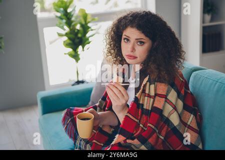 Foto ritratto di una giovane ragazza dai capelli ricci seduta sul comodo divano, che guarda il termometro e beve il tè trattato all'interno Foto Stock