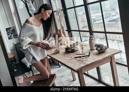 Colazione deliziosa. Vista dall'alto di una giovane donna attraente che versa cereali e sorride mentre si trova vicino alla finestra di casa Foto Stock