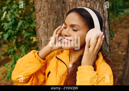 Una giovane donna afroamericana sorride mentre cammina attraverso una foresta autunnale, ascoltando la musica con gioia. Foto Stock