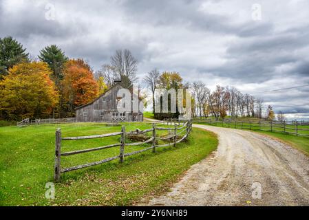 Vecchio fienile di legno lungo una strada di ghiaia che attraversa boschi al culmine dei colori autunnali in una giornata nuvolosa Foto Stock