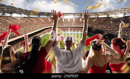 Vivace scenario di stadio di calcio pieno di tifosi che sventolano bandiere rosse e coppe, con coriandoli luminosi che cadono intorno a loro, mentre il sole splende sul campo. Foto Stock