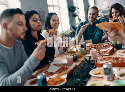 Migliori amici. Gruppo di giovani in abbigliamento casual che mangiano pizza e sorridono mentre si fa una cena all'interno Foto Stock