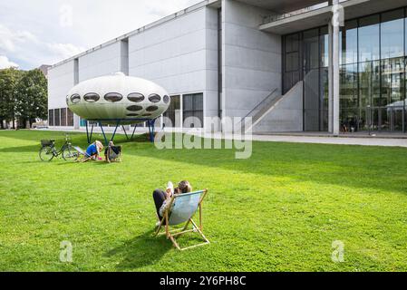 Persone sedute su sdraio sul prato di fronte alla Pinakothek der moderne e all'installazione artistica futuro House di Matti Suuronen, Monaco di Baviera Foto Stock