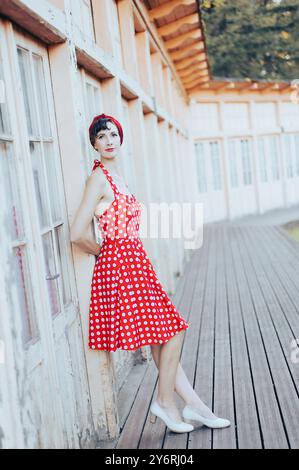 Una giovane donna con un abito a pois rosso si appoggia su un edificio rustico con pannelli di legno, che mostra uno stile vintage alla moda. Foto Stock