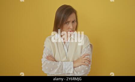 Una donna matura scontenta con i capelli corti regge le braccia incrociate su un solido sfondo giallo, esprimendo insoddisfazione. Foto Stock