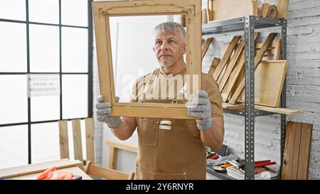 Bell'uomo anziano che tiene il telaio in legno in un'officina di falegnameria ben attrezzata Foto Stock