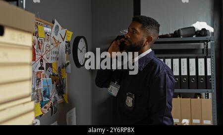 Detective afroamericano che analizza le prove in un ufficio della stazione di polizia, parla al telefono con un'espressione seria. Foto Stock