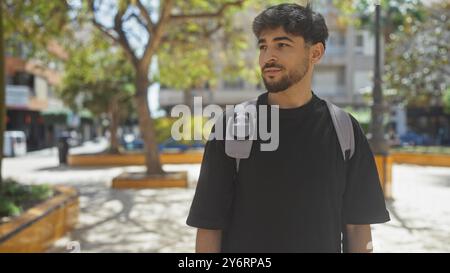 Bel giovane arabo adulto con la barba, vestito in modo informale con una borsa a tracolla, in posizione contemplativa in un parco urbano. Foto Stock
