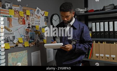 Detective afroamericano che prende appunti in un ufficio con una scheda delle prove, che indica la scena dell'indagine. Foto Stock