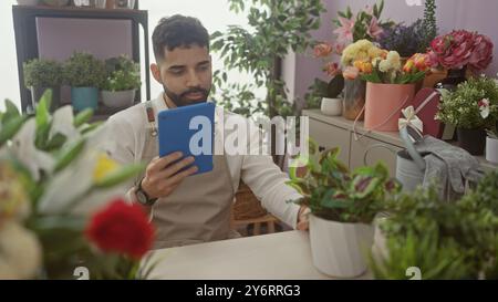 Un giovane ispanico con la barba esamina una tavoletta in un negozio di fiori circondato da varie piante in vaso. Foto Stock