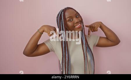 Donna africana sicura di sé con trecce che puntano a se stessa su uno sfondo rosa isolato, incarnando bellezza e positività. Foto Stock
