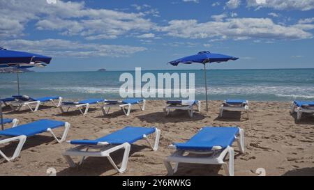 Lettini e ombrelloni blu su una spiaggia di sabbia che si affaccia sul mare calmo sotto un cielo nuvoloso, raffigurando una tipica scena di vacanza estiva. Foto Stock