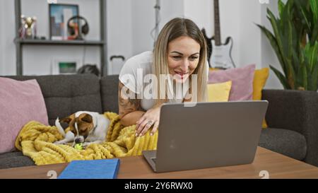 Una giovane donna con un tatuaggio sorride mentre lavora sul suo portatile a casa, con il suo cane che gioca nelle vicinanze. Foto Stock