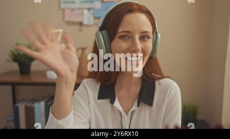 Una giovane donna sorridente con le cuffie gode di una videochiamata nel suo accogliente ufficio domestico. Foto Stock